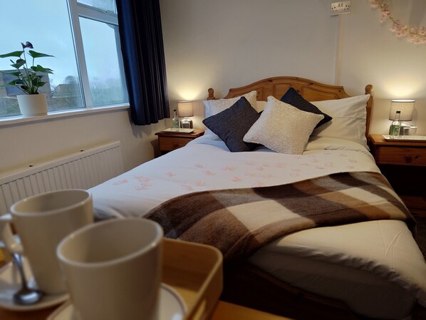 A neatly made bed with a floral bedspread, two bedside lamps, a potted plant, and a window with curtains. - Windsor Local Hotel (Johnston)
