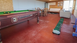 A pool table with chairs, a sofa, and a billiards cue rack in a room with wooden walls and a red floor. - Cabin 9 Inis Beag (Gortahork)