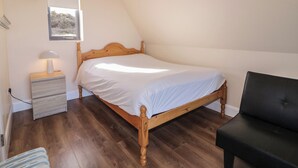 A bedroom with a wooden bed, a nightstand with a lamp, a chair, and a window with a view of the outdoors. - Cabin 9 Inis Beag (Gortahork)