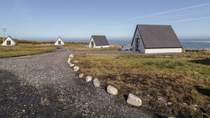 A coastal area with three small, white, single-story buildings with dark roofs, arranged in a row along a gravel path. - Cabin 9 Inis Beag (Gortahork)