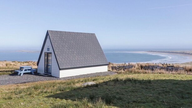 A small, white, single-story building with a sloped roof, situated on a grassy hill overlooking a beach and ocean. - Cabin 10 Inis Bo Finne (Gortahork)