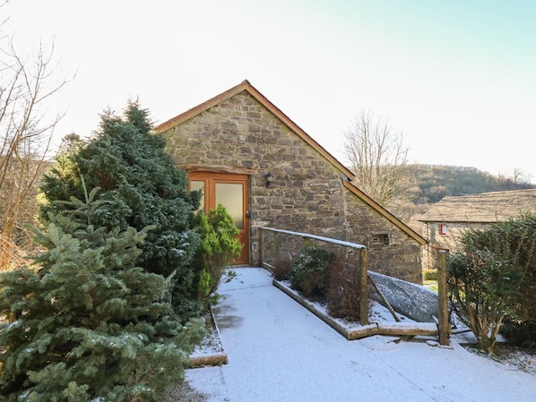 Un edificio de piedra con una puerta de madera, un sendero cubierto de nieve y un pequeño árbol en primer plano.