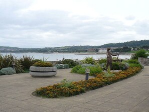 A landscaped area with a statue, a stone planter, and a view of a body of water and distant buildings. - Ocean View Apartment, Rhos-On-Sea (Colwyn Bay)