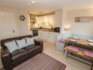 A compact living space with a kitchenette, a sofa, a dining table with benches, and a clock on the wall. - Clickety-Clack Cottage, Whitby (Whitby)