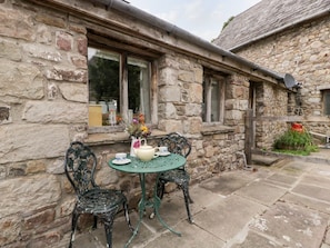 Ein Steinhaus mit einer kleinen Terrasse, auf der ein Tisch und zwei Stühle stehen, sowie ein Fenster und eine Satellitenschüssel.