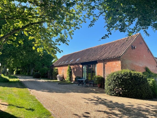 A brick building with a sloped roof, a gravel driveway, and a small patio area with tables and chairs. - Pear Tree, Wood Norton (Dereham)
