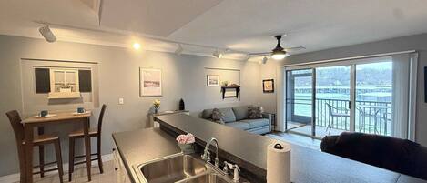 A modern kitchen with a large island, a dining area with chairs, a sofa, and a view of a balcony with outdoor seating.