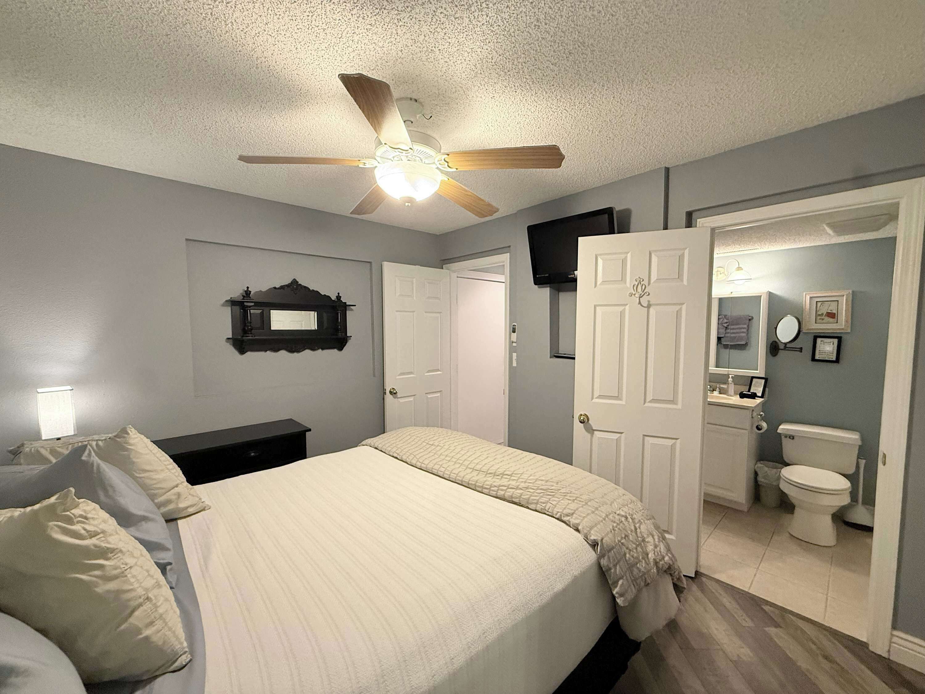A bedroom with a bed, a ceiling fan, a door, a mirror, and a bathroom with a toilet and sink.