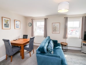 A room with a dining table, chairs, a sofa, a lamp, a clock, and a window with curtains. - Rails End, Whitby, North Yorkshire (Whitby)