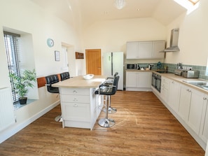 A modern kitchen with white cabinetry, a central island with bar stools, a microwave, oven, and a sink. - Ty Cuddfan, Llangoed (Beaumaris)