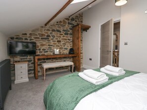 A bedroom with a stone wall, a wooden desk, a television, a bench, a bed with green and white bedding, and a door. - Howgill House, Sedbergh, Wi-Fi (Sedbergh)