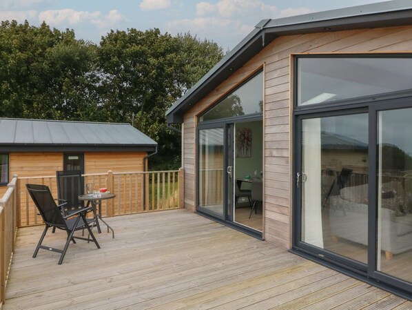 A wooden deck with a table and chairs, a sliding glass door, and a small building in the background.