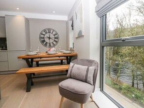 A modern kitchen with a wooden table and bench, a large wall clock, and a view of a river through a window. - Brimstone, Backbarrow, Cumrbia (Ulverston)