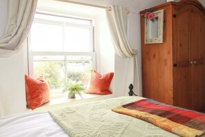 Une chambre à coucher avec un lit, une fenêtre avec des rideaux, une armoire en bois et une petite plante posée sur le rebord de la fenêtre.