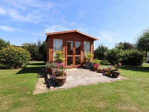 A small wooden cabin with a patio, surrounded by potted plants and a well-maintained lawn. - The Flat At Brwyn, Brynford (BRYNFORD)