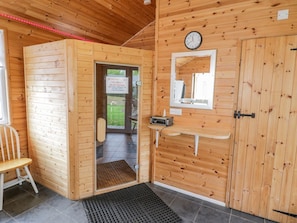 Eine Holzhütte mit Holztür, einer Uhr, einem kleinen Regal mit einem Radio, einem Stuhl und einem Fenster.