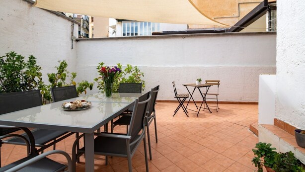 Una terrazza con tavolo in vetro, sedie e un piccolo tavolino con una sedia. Sono presenti piante in vaso e una grande vela ombreggiante.