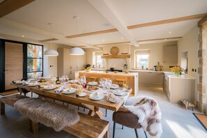 A modern kitchen with a central island, wooden beams, pendant lighting, and a dining area with a long table set for a meal. - The Malt Kiln is nr Harrogate. (Harrogate)