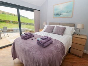Ein Schlafzimmer mit Bett, zwei Kissen, einem Nachttisch, einer Lampe und Blick auf eine grüne Landschaft durch ein großes Fenster.