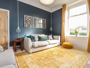 A room with a dark blue wall, a white sofa, a yellow patterned rug, and a window with curtains. - Castlewood Cottage, Skipton (Skipton)