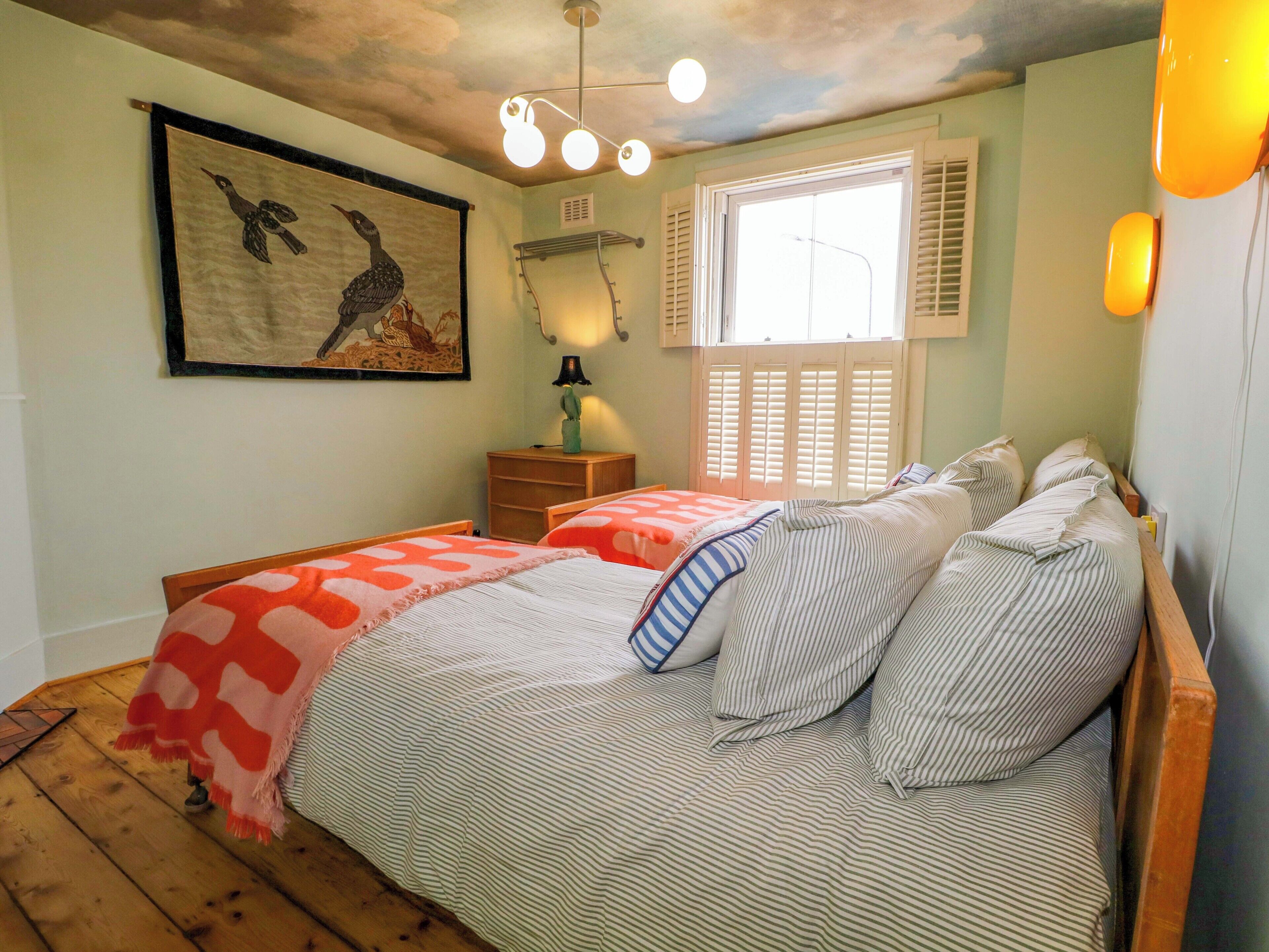 A bedroom with two beds, a window with shutters, a ceiling light fixture, and a wall-mounted lamp.