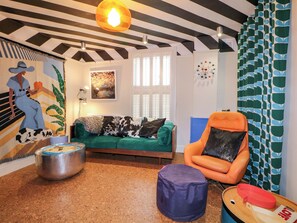 Um quarto com teto listrado, sofá verde, cadeira laranja, um banquinho redondo e um pote de metal no chão.