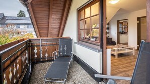 Un balcon doté d’une balustrade en bois, d’un fauteuil noir, d’une table blanche et offrant une vue sur une maison équipée de panneaux solaires.