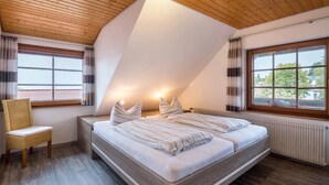 Une chambre avec un plafond en pente, un lit avec une literie à rayures, une chaise et deux fenêtres avec des rideaux.