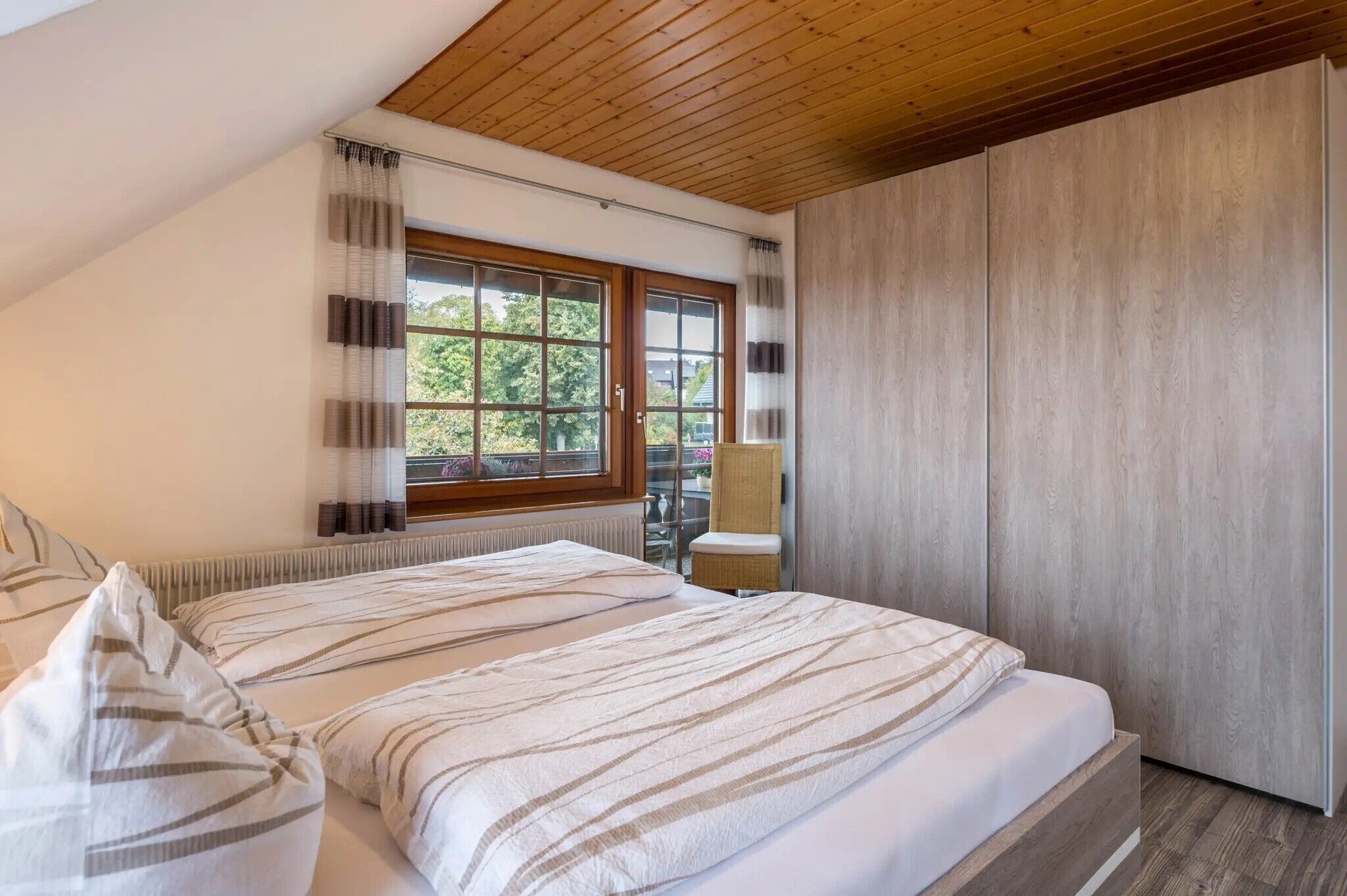 A bedroom with a sloped ceiling, a large window, a bed with striped bedding, a chair, and a wardrobe.