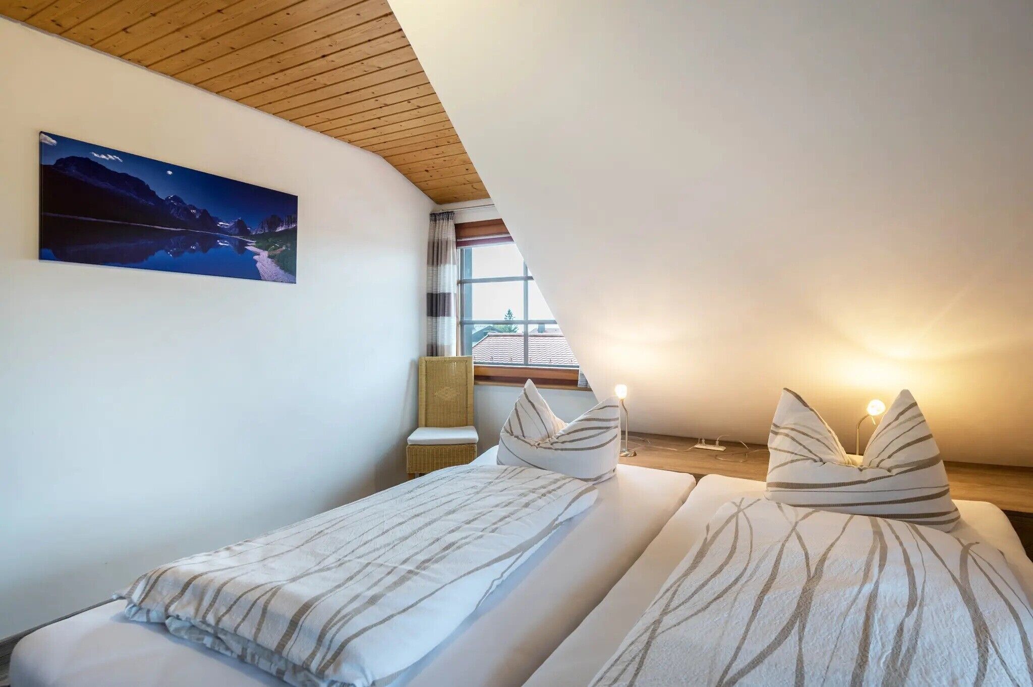 Un dormitorio con cama, almohadas, una mesita de noche, lámpara, ventana y un cuadro colgado en la pared.