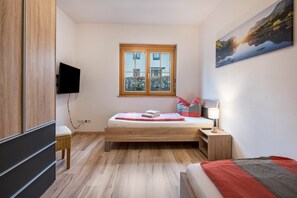 Une chambre à coucher avec un lit, une table de chevet avec une lampe, un téléviseur fixé au mur et une fenêtre donnant sur un immeuble et des arbres.