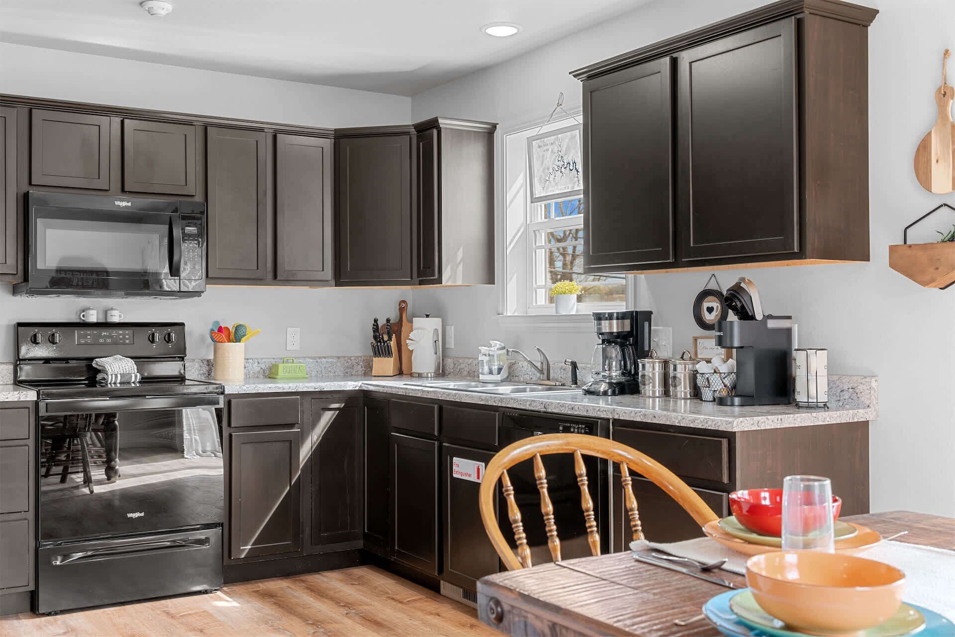 A modern kitchen with dark cabinetry, a built-in microwave, stove, and dishwasher. There's a dining area with a wooden table and chairs.