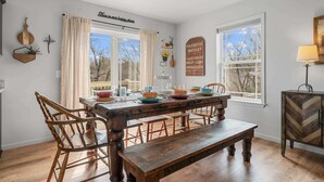A dining area with a wooden table, chairs, and a bench. There are two windows with curtains, a wall-mounted sign, and a decorative wall hanging. - Trolly Inn Hot Tub, Walkable (Branson)