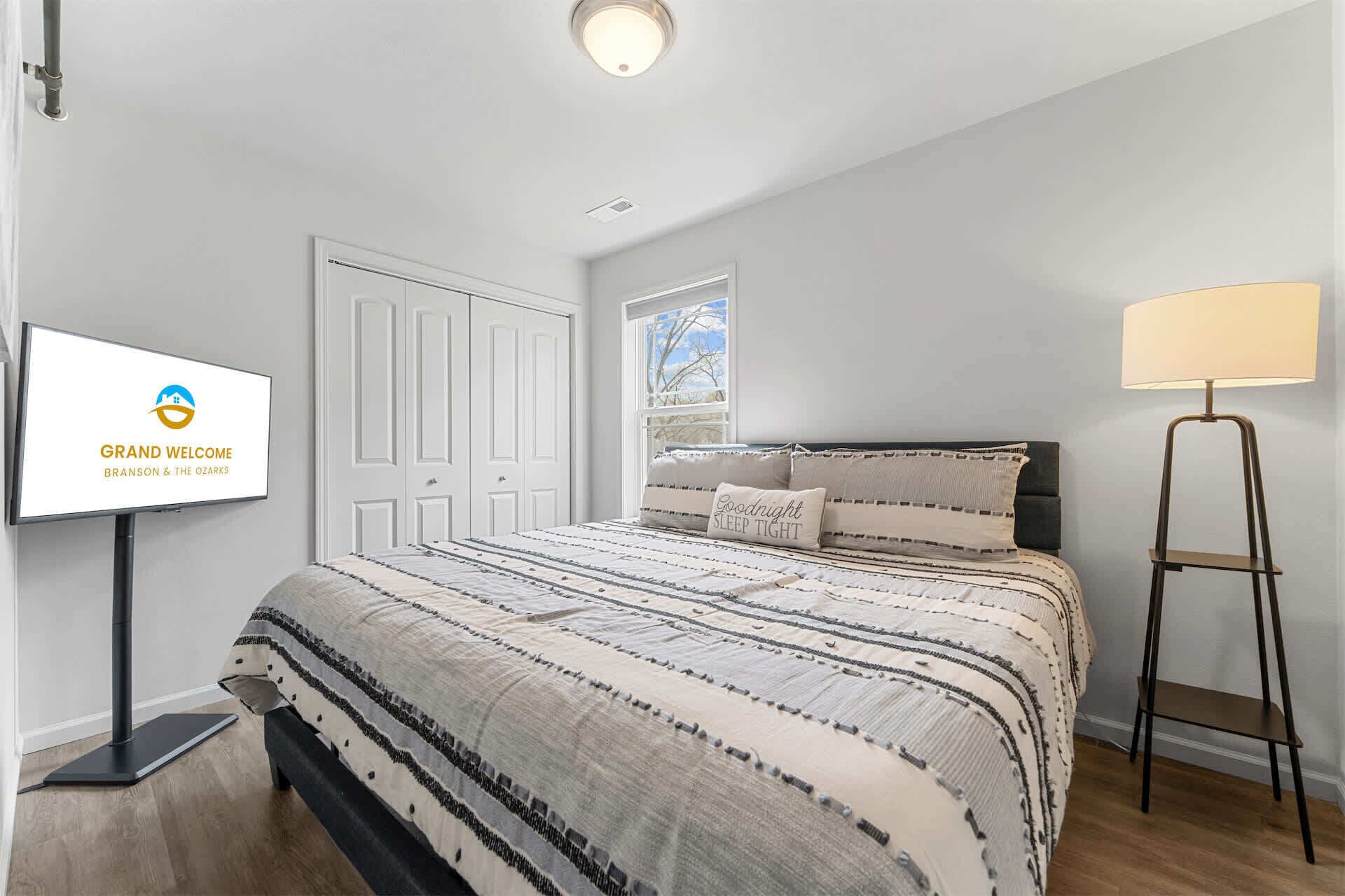 A bedroom with a bed, a standing lamp, a TV, and a window.