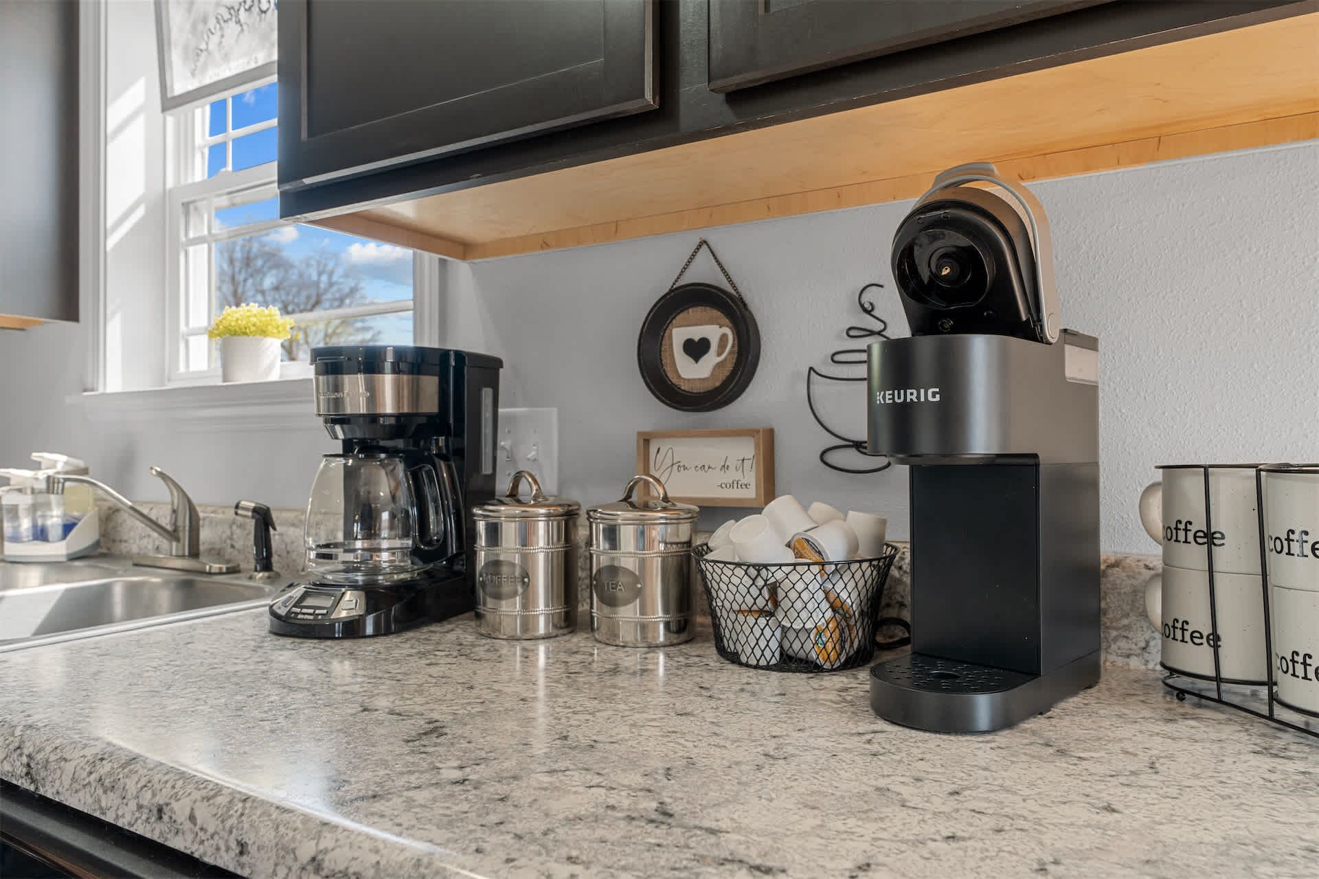 A kitchen with a coffee maker, tea pot, and coffee cups on the counter.