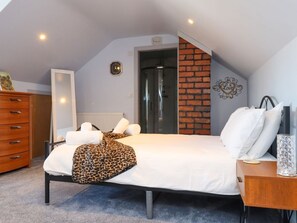 A bedroom with a bed, a wooden dresser, a mirror, a brick accent wall, and a door leading to another room. - Afallon, Llanerchymedd (Llannerch-y-Medd)