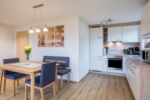 Eine moderne Küche mit weißen Schränken, einem eingebauten Backofen und einer Mikrowelle. Es gibt eine Essecke mit einem Holztisch und blauen Stühlen.
