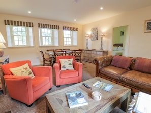 A living room with two red armchairs, a brown leather sofa, a wooden coffee table with books and a remote control, a dining area with a table and chairs, and a green wall with a clock. - Kilnmark Farmhouse in Moniaive (Thornhill)
