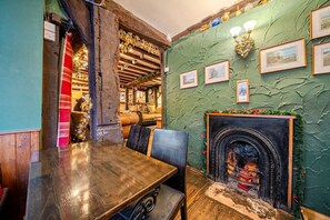 Un interior rústico con una mesa de madera, sillas, una chimenea y una pared verde con cuadros enmarcados.
