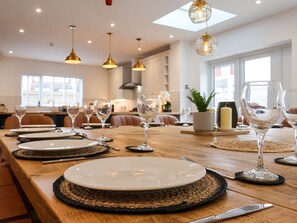 A dining area with a long wooden table set for a meal, overhead pendant lighting, and a view of the kitchen in the background.