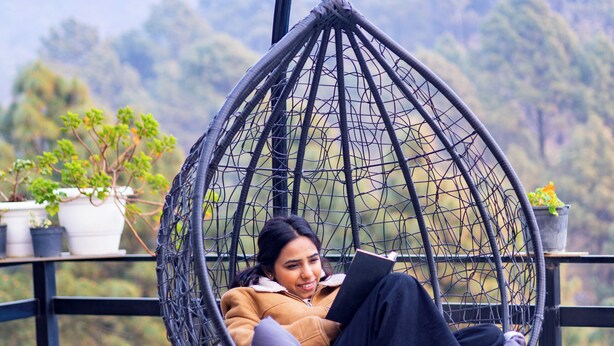 A person in a hanging chair on a balcony with potted plants and a view of hills. - ELIVAAS The Nakshatra Serene 3BHK Villa (Sukha)