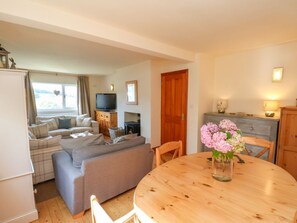 A living room with a sofa, a television, a wooden dining table with a vase of flowers, and a kitchen area with cabinets and a refrigerator. - Wayside, North Yorkshire 3 bed (Whitby)
