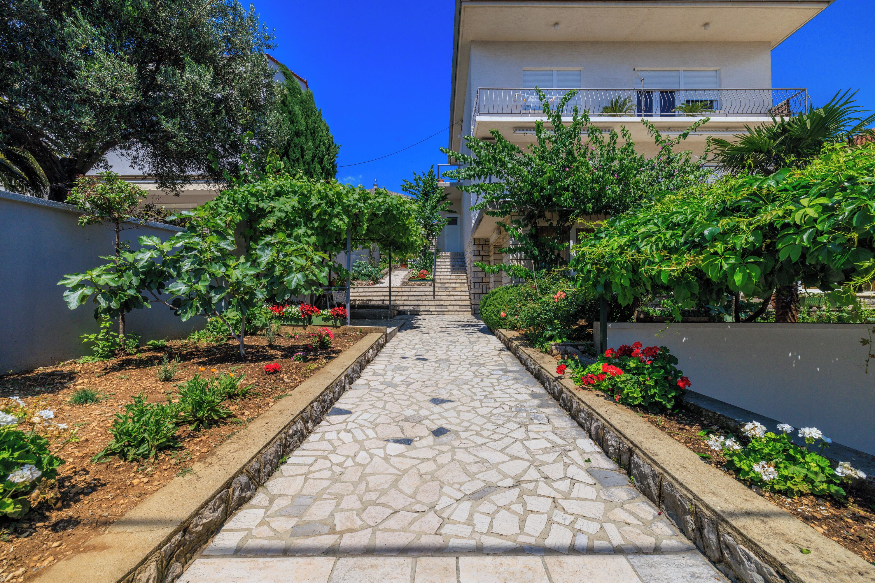 A stone pathway leading to a building with a balcony, surrounded by greenery and plants.