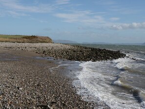 A rocky beach with waves, a grassy area, and distant hills. - Beudy Bach, Llangoed (Beaumaris)
