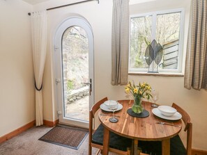 A dining area with a round wooden table, chairs, a vase with flowers, a window with curtains, and a door with a view of the outside. - Mill Cottage, Buckden (Skipton)