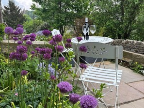 A garden setting with a white metal chair, a table with a bottle and glasses, and purple flowers. - Angel's Cottage, Tideswell. TV (Buxton)