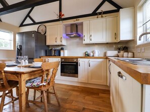A kitchen with white cabinets, a stainless steel refrigerator, a wooden dining table with chairs, and a window with blinds. - White Hart Stables, Catsfield (Battle)