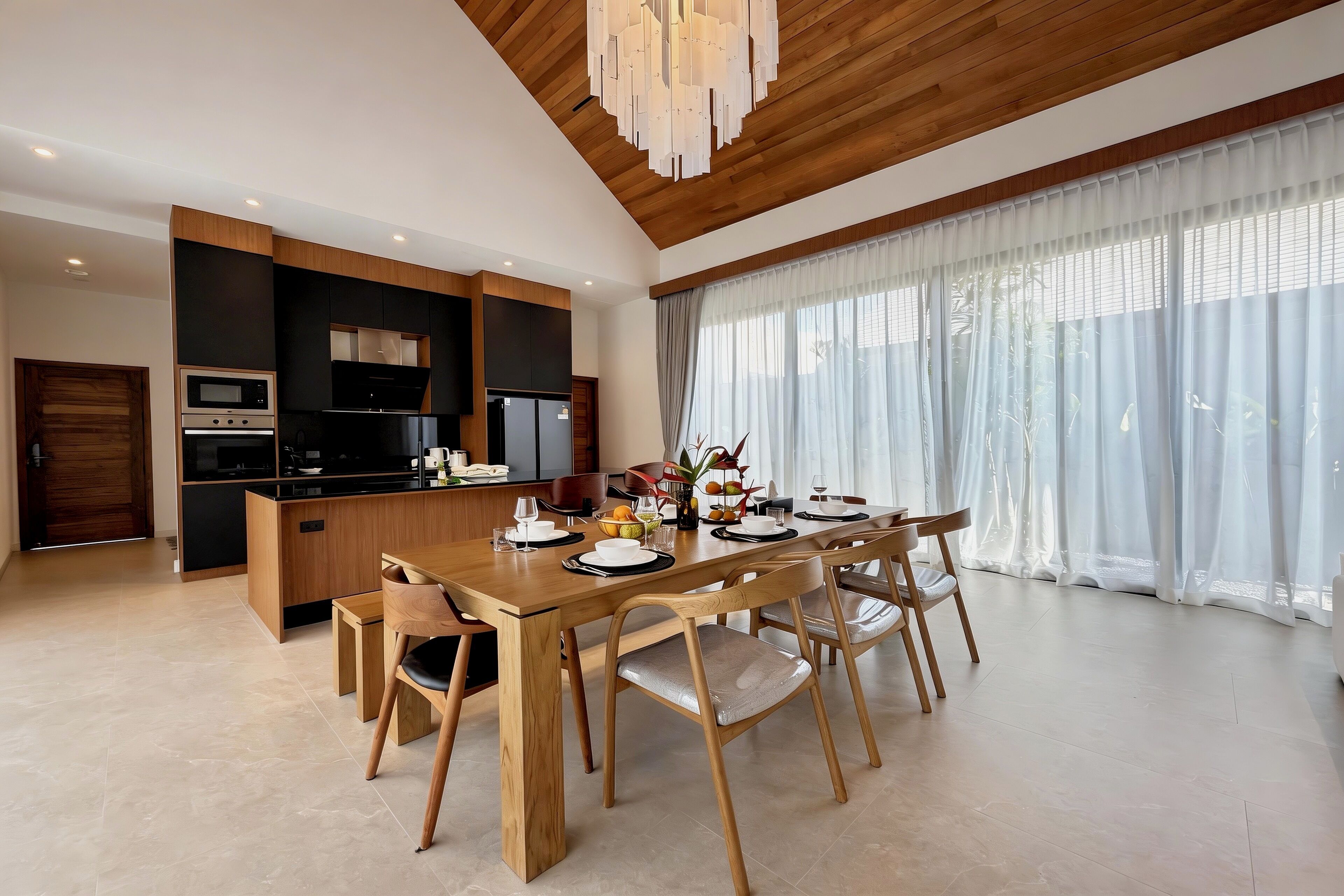 Una cocina moderna con electrodomésticos integrados, un comedor con mesa y sillas de madera y un amplio ventanal con cortinas de gasa.