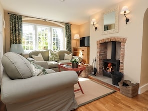 A living room with a sofa, a fireplace, a television, and a window with curtains. - The Alms House East is in Forthampton (Gloucester)