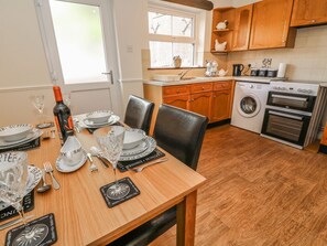 A dining area with a table set for a meal, a washing machine, and a kitchen with wooden cabinets and appliances. - Charming Croft Cottage Retreat (Skipton)